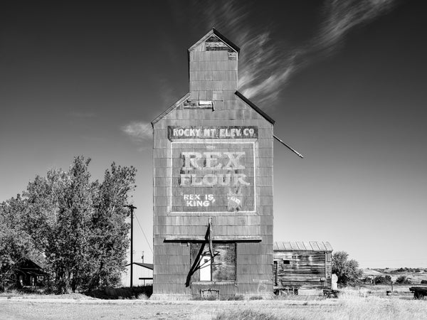 Black and white photograph of an old grain elevator standing in a very small Montana town The fading painted sign says, "Rocky Mountain Elevator Company. Rex Flour. Rex is King.