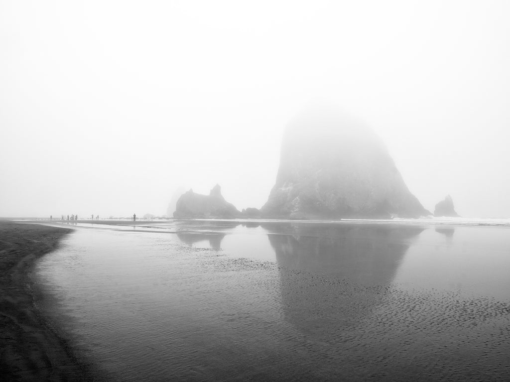 Black and white photograph of Haystack Rock, Oregon's iconic landmark on the coast at Cannon Beach, cloaked in a layer of dense morning fog. People on the shoreline give a sense of scale of the massive rock.