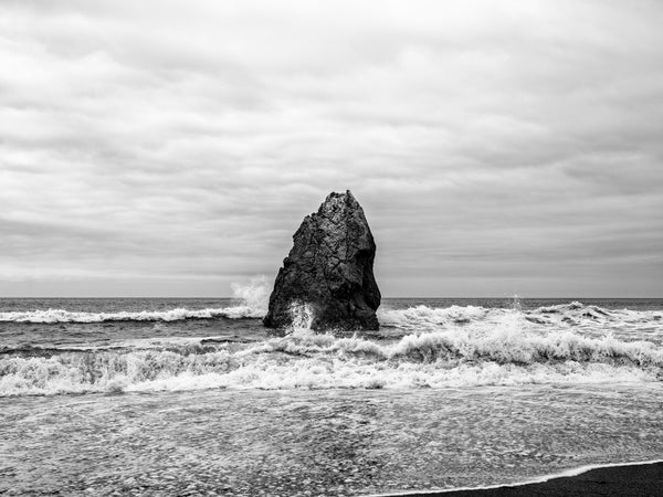 Black and white photograph of a giant rock formation standing like a sentinel amidst tumultuous Pacific Ocean surf on the Oregon Coast.