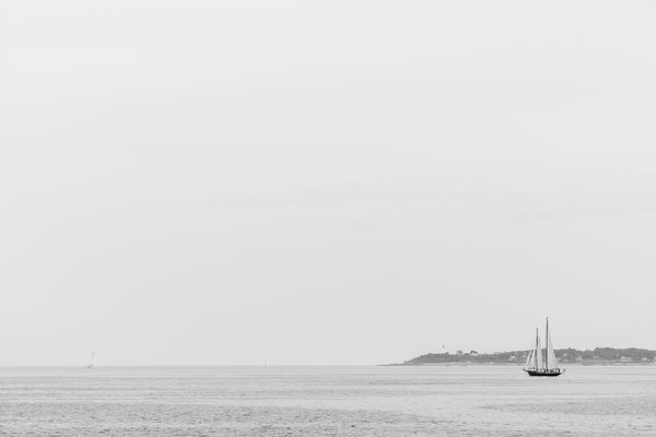 Black and white photograph of sail boats cutting through sun-washed haze of the New England coast with a lighthouse visible in the distance.