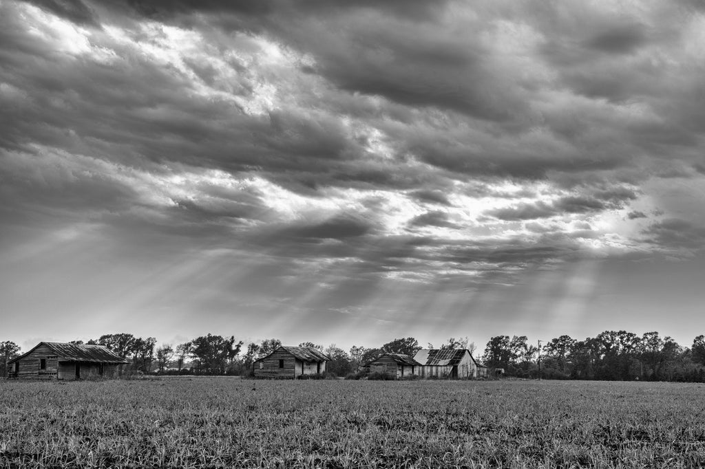 Black and white photograph of rays of sunset bursting through storm clouds over a collection of abandoned old farm shacks in the American South.