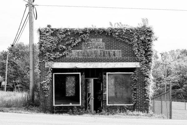 Black and white photograph of the historic abandoned Bank of Centertown, Tennessee, built 1919. A large sign over the front doors includes the name and date, surrounded on the sides by encroaching ivy.