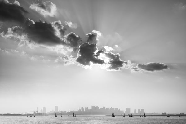 Black and white photograph of the distant skyline of Boston with sailboats in the harbor and a burst of sun rays behind clouds at sunset.