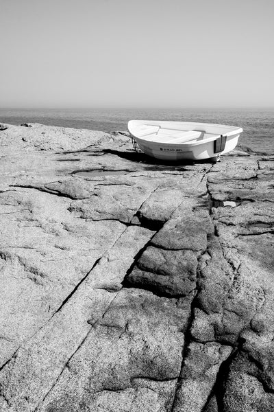 Black and white photograph of a small boat perched on the rocky sea coast of Maine overlooking the vast Atlantic Ocean.