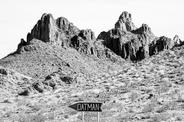 Black and white photograph of a handmade sign that points to the community of Oatman, on an older alignment of Route 66 in the rugged Arizona mountains.