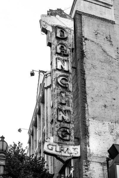 Black and white photograph of a rusty and crusty old neon sign that says "Dancing Girls," seen on a gritty street in downtown Los Angeles. 