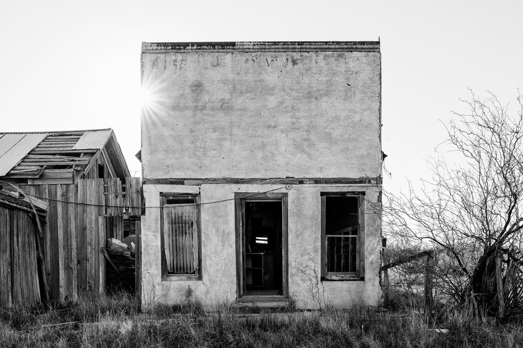 Black and white photograph of the sun rising behind an abandoned old store with doors and windows missing in the near-ghost town of Toyah in the desert of West Texas.