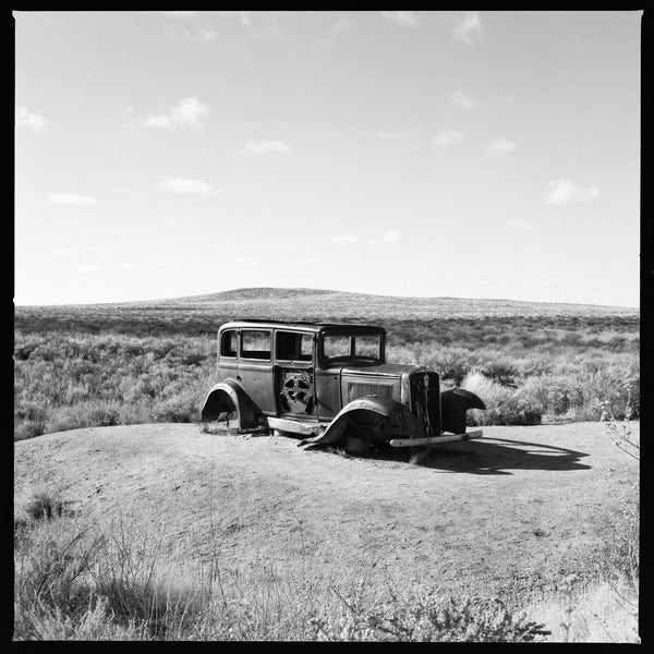 Black and white photograph of a rusty 1932 Studebaker positioned on a historic alignment of Route 66 in the Petrified Forest National Park. This print includes a narrow black portion of the film edge as shown.