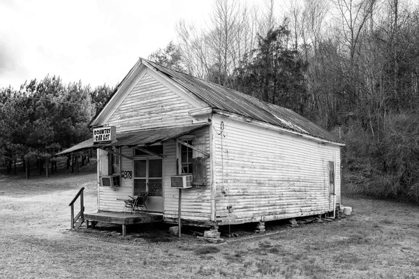 Black and white photograph of an abandoned old general store building that ended its useful existence as a used car lot along a back road in the rural American South.