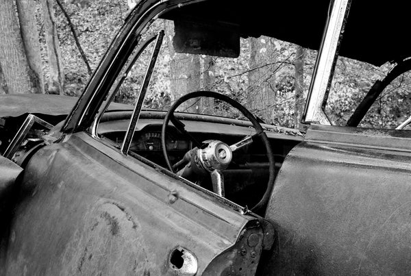Black and white photograph of a classic American car slammed into a tree in the forest and abandoned decades ago.