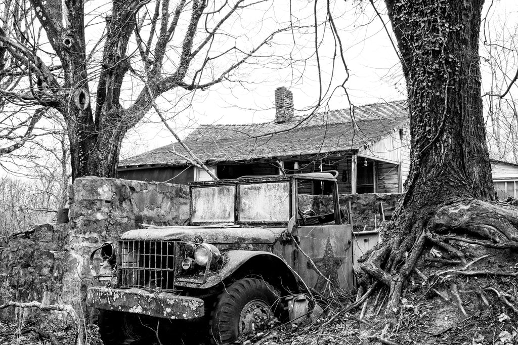 Black and white photograph of an abandoned old house with a big antique military truck with a star on its side parked in the front yard.