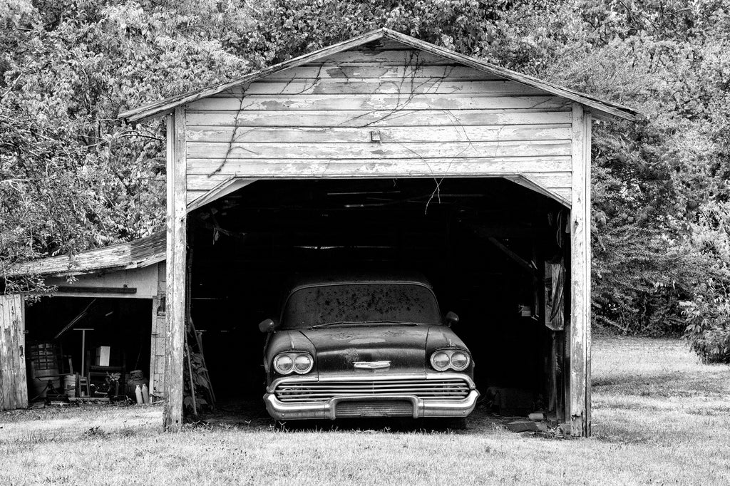 Black and white photograph of a dusty black classic American car left parked in an old wooden garage.