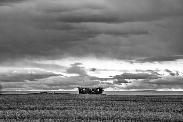 Black and white photograph of the remains of a collapsed, isolated old wooden farmhouse until stormy skies on the vast high prairie farmland of Montana.