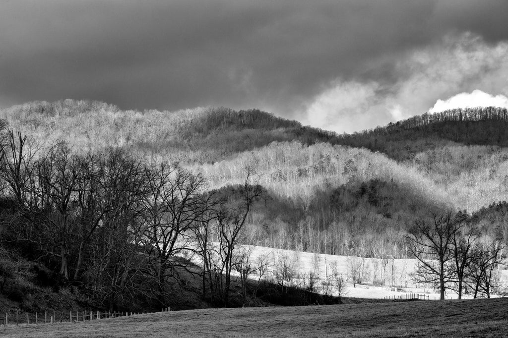 Dramatic black and white landscape photograph with dark storm clouds casting dark shadows over the rolling hills of Virginia.