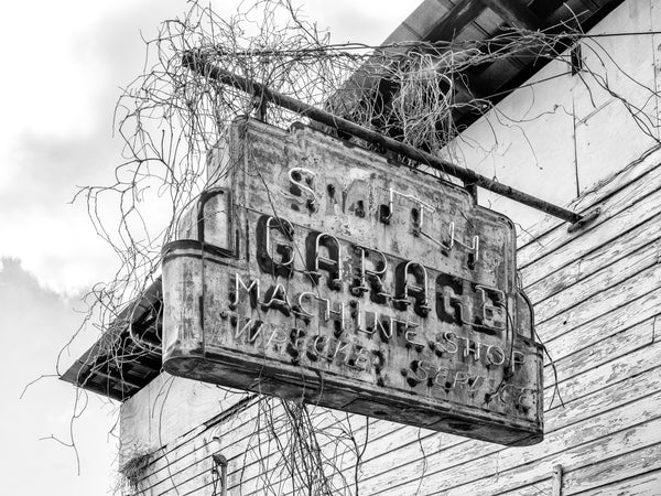 Black and white photograph of a rusty, overgrown vintage neon sign for "Smith Garage, Machine Shop, and Wrecker Service," found in a small town in the American South.