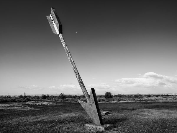 Black and white photograph of the moon behind the last arrow standing at the old Twin Arrows Trading Post, now abandoned in ruins on old Route 66 in Arizona.
