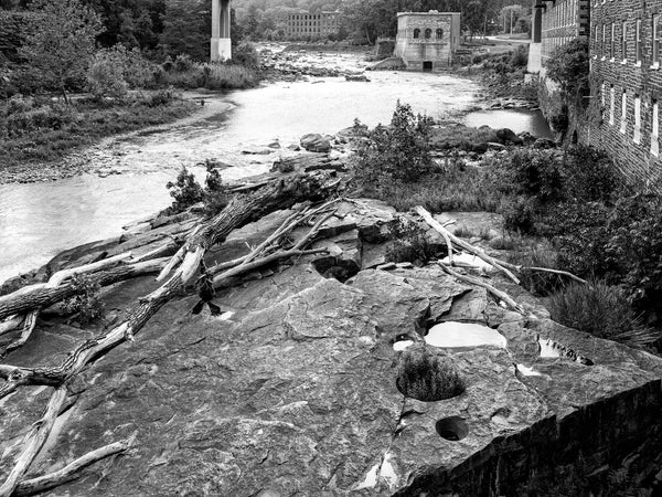 Black and white landscape photograph of a rocky riverbed with fallen logs, water, industrial structures, and a brick building along the right side.