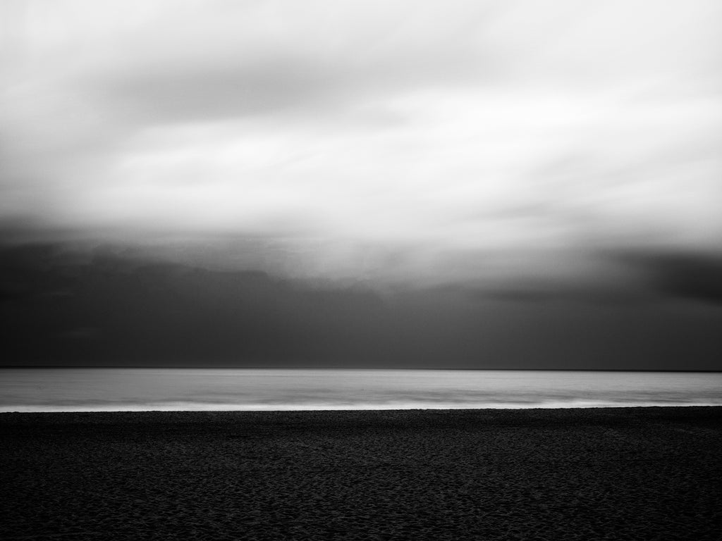 Black and white long-exposure photograph of the New England coastline with deep, dark storm clouds approaching over the silvery Atlantic Ocean.