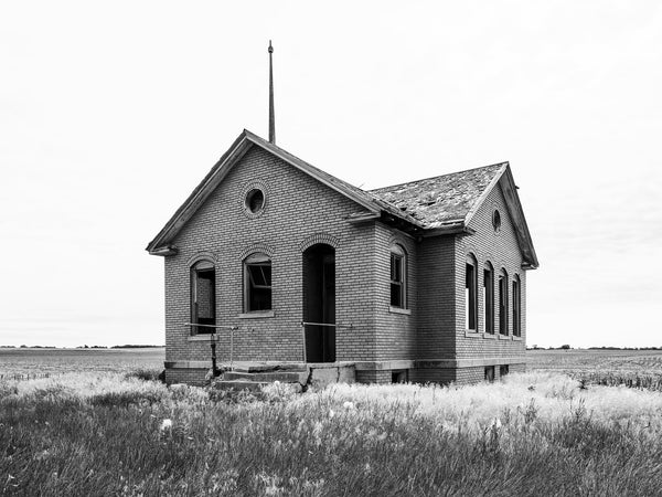 Black and white photograph of a red brick one-room schoolhouse built in 1920 that stands abandoned along an unpaved country road in the wide open Kansas prairie.
