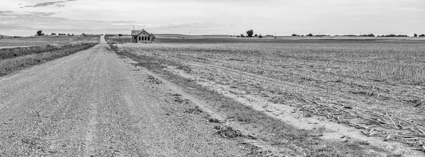 Black and white panoramic landscape photograph of a red brick one-room schoolhouse built in 1920 that still stands stoically along an unpaved country road in the wide open Kansas prairie.