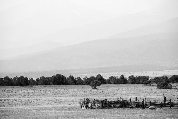 Black and white fine art landscape photograph of hazy Colorado mountains in the background, with an abandoned collapsing corral in the foreground.