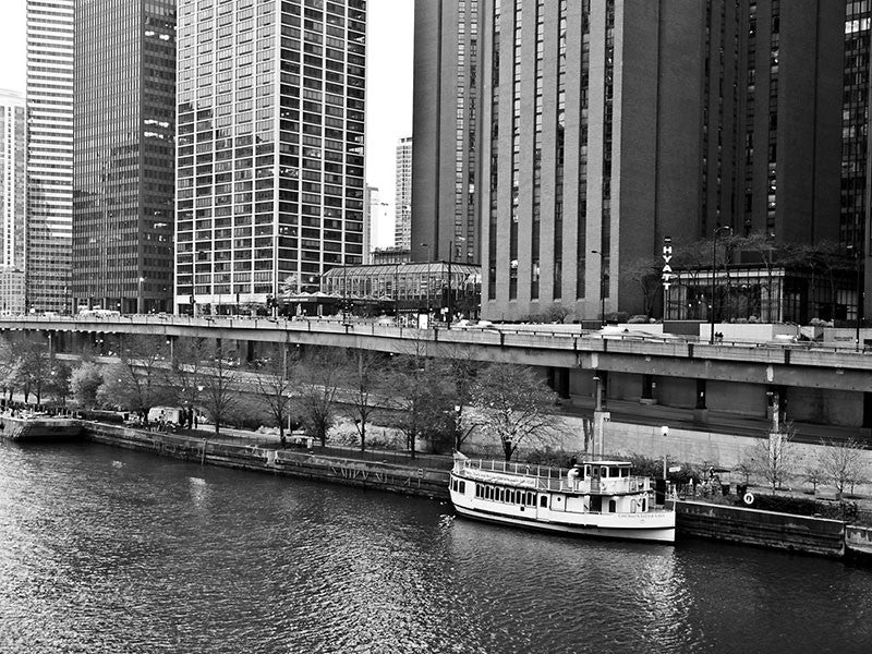 Black and white photograph of a boat on the Chicago River, dwarfed by skyscrapers in downtown Chicago.