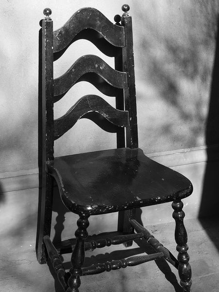 Black and white photograph of an old wooden black chair situated in a sunny location near and adobe wall in Santa Fe, New Mexico.