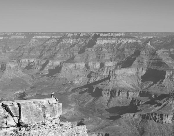 Black and white landscape photograph of the south rim of the Grand Canyon just after sunrise, with a guy sitting on the point of a precipice on the edge.