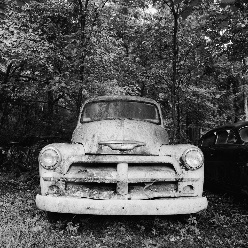 Black and white photograph of a pale-colored antique pickup truck abandoned in a dark forest. 