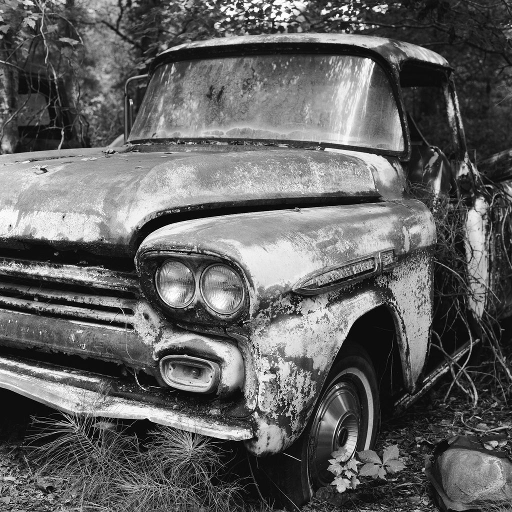 Black and white photograph of a rusty antique Chevrolet pickup truck in a wooded junkyard in the American South.
