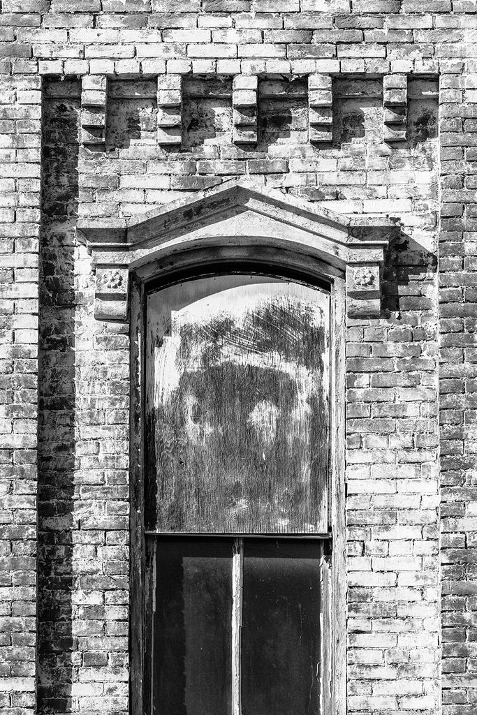 Black and white photograph of a window on an abandoned brick building with a stone pediment and paint peeling from its bricks.