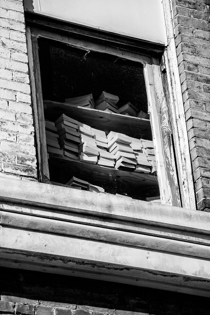 Black and white photograph of a window on the second floor of an abandoned building that shows stacks of paperback books on shelves.