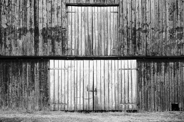 Black and white photograph of barn doors on the first and second levels of a big old weather-beaten red barn. This photograph is composed flatly, allowing the rectangles and vertical lines to create an almost abstract architectural image.
