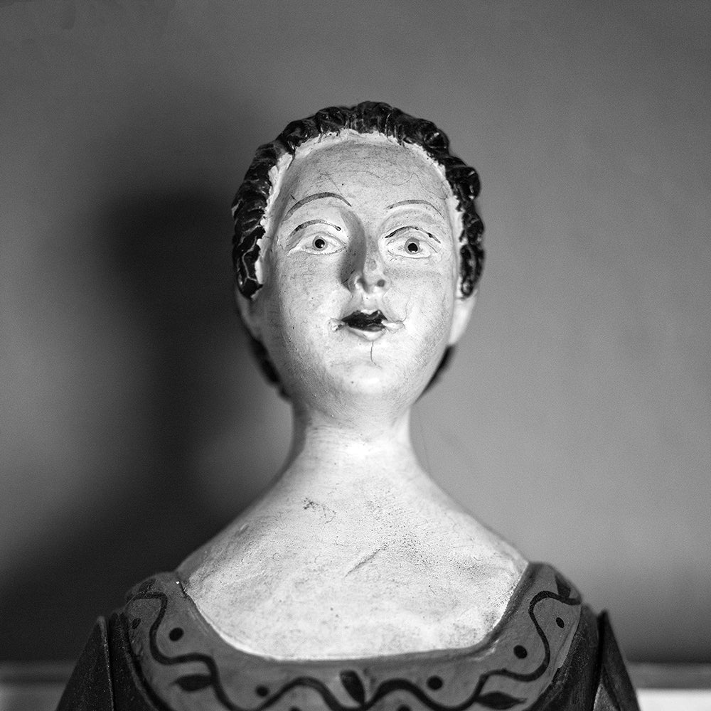 Black and white photograph of an antique wooden doll with hand-painted features found in an old house, photographed portrait style. (Square format)