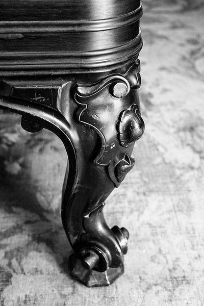 Black and white photograph of an ornate table leg standing on a threadbare rug inside a big old house.