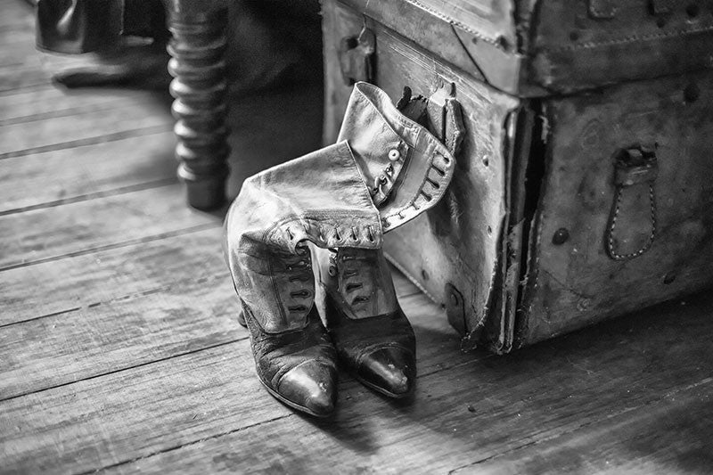 Black and white photograph of a pair of old Victorian-era "granny boots" displayed beside a tattered leather trunk, found in the attic of an old house.
