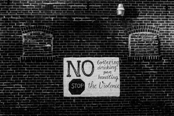 Black and white photograph of a hand-painted sign on a brick wall in North St. Louis, Missouri. The text reads "No loitering drinking panhandling. Stop the violence."