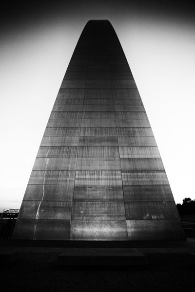 Black and white photograph of the famous St. Louis Gateway Arch at sunset, seen from the point of view of the base, looking up its steep metallic slope. 
