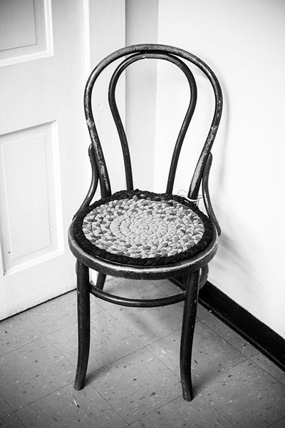 Black and white photograph of an antique chair with homemade cushion sitting in a corner in a big old house.