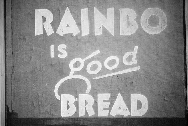 Black and white photograph of a vintage ad for Rainbo Bread, as seen on a screen door. The ad reads, "Rainbo is good bread."