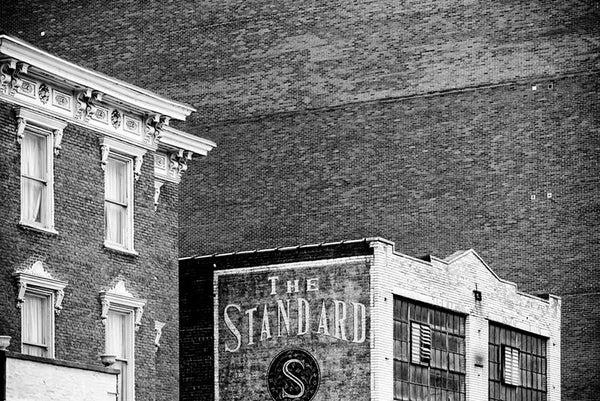 Black and white architectural photograph of brick buildings, including a vast brick wall, in downtown Nashville. The side of a smaller building displays a painted sign for The Standard.