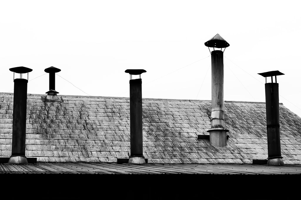 Moody black and white photograph of a tin roof with five chimney pipes rising up at various heights creating an interesting visual rhythm. 