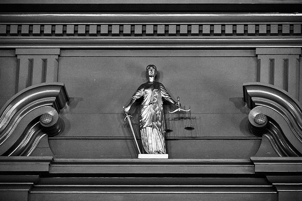 Black and white photograph of a small, golden "Justice is Blind" statue on the interior of Independence Hall in Philadelphia.