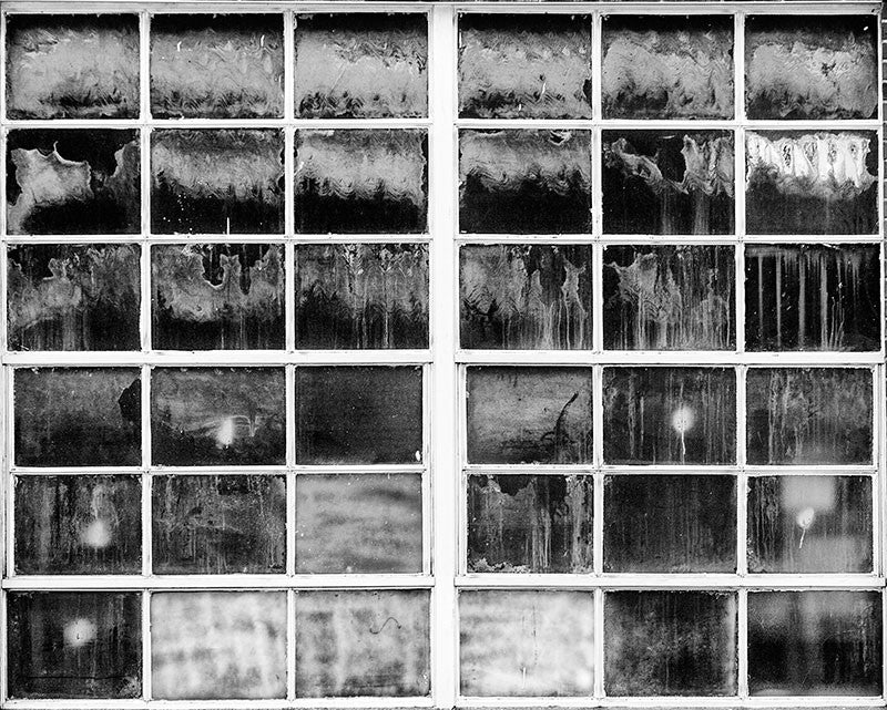 Black and white photograph of a grid of industrial windows with myriad textures, splashes, stains and tones. Each pane of glass looks like an individual work of art.