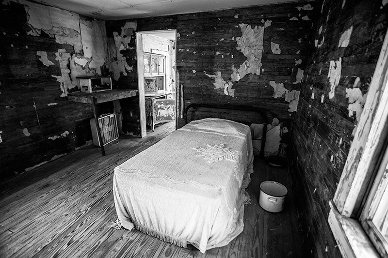 Black and white photograph inside the bedroom of the late blues musician Sleepy John Estes. Here we see his bed, covered by a bedspread, siting on bare wood floors and surrounded by wooden walls with small scraps of remaining original wallpaper. Estes small house in Brownsville, Tennessee has been preserved and relocated to a visitors center near the schoolhouse of Tina Turner, who also came from Brownsville.