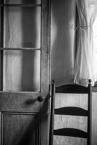 Black and white photograph of the inside of an old Cajun house in Lafayette Louisiana, with a French-style door, a simple wooden chair, and a sheer curtain.