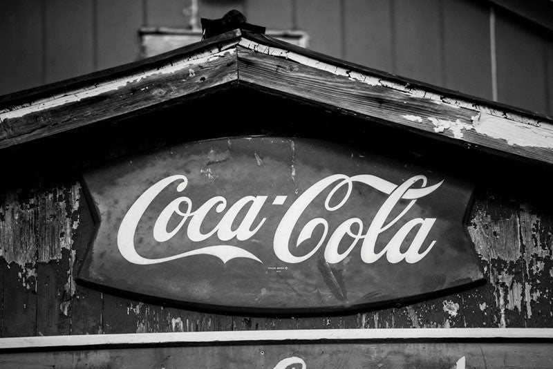 Black and white photograph of a vintage Coca Cola brand sign on a very old building with peeling paint in a small southern town.