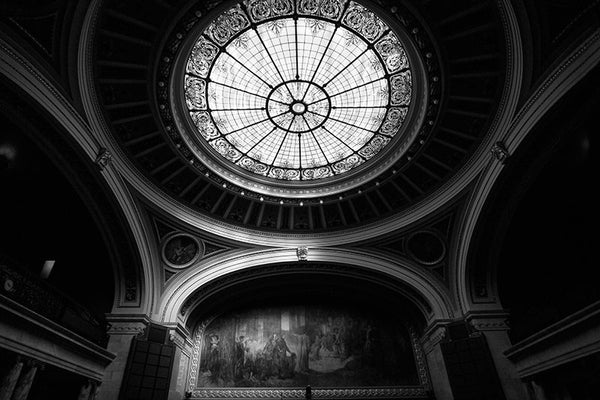 Black and white natural light photograph of a highly ornate assembly room inside the Wisconsin state capitol in Madison, Wisconsin. Because the assembly was not in session, the lights were off in the room, with just natural light from the decorative overhead skylight to illuminate the massive mural at the head of the room.
