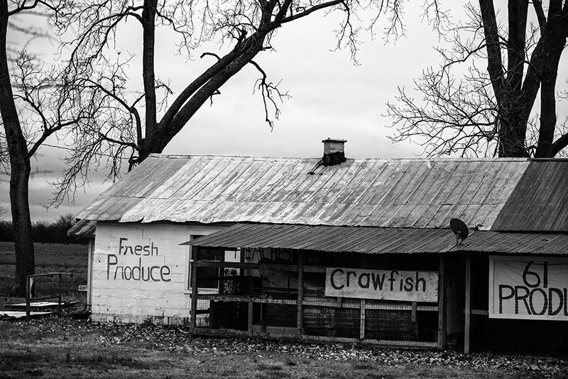 Black and white photograph of a roadside farm fresh produce stand near Tunica, Mississippi. On the barn are hand-painted signs that say "Fresh Produce," and "Crawfish."