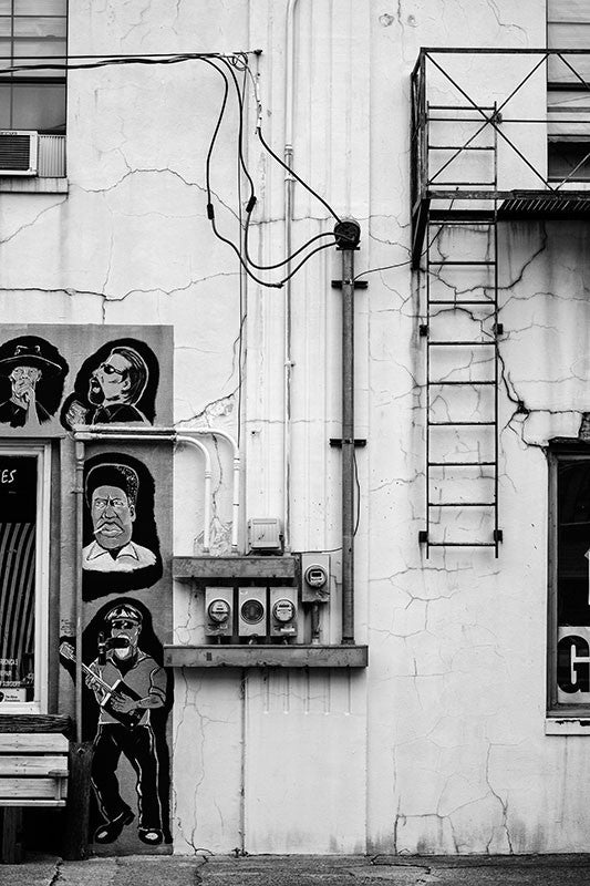 Black and white photograph of a street scene in downtown Clarksdale, Mississippi, with paintings of bluesmen, utility meters, and a fire escape ladder.